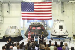 American Flag hanging above three space capsules. Seated crowd being addressed by NASA speakers. Airborne Systems. Design, development, and manufacture of space parachute & inflatable systems. Military-grade deceleration, airbag landing, aerospace recovery, personnel & cargo delivery parachute systems.