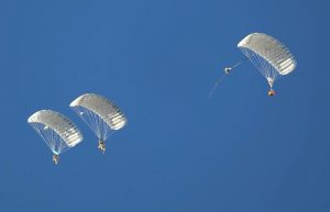 Airborne Systems Combo Drop MicroFly II Army Cargo delivery system. JPADS / GPADS: Guided Precision Aerial Delivery System. Use with any Airborne Systems Ram Air Canopy. Two troop jumpers with deployed canopy and cargo parachute blue sky.