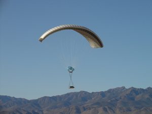 Airborne Systems DragonFly Army Cargo Delivery Parachute System flying with cargo. JPADS 10K System of Choice. Eliptical canopy carries loads up to 10,000 lbs. Max altitude 24,500 ft. View of canopy and payload from the side with mountains behind. 10k