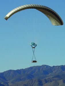 Airborne Systems DragonFly Army Cargo Delivery Parachute System flying with cargo. JPADS 10K System of Choice. Eliptical canopy carries loads up to 10,000 lbs. Max altitude 24,500 ft. View of canopy and payload from the side with mountains behind.