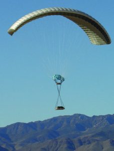 Airborne Systems DragonFly Army Cargo Delivery Parachute System. JPADS 10K System of Choice. Eliptical canopy carries loads up to 10,000 lbs. Max altitude 24,500 ft. Canopy and blue sky, mountains.