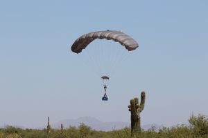 Airborne Systems. FC Mini Army cargo delivery parachute system. JPADS / GPADS: Guided Precision Aerial Delivery System. Carries 200-500 lbs. Max deployment altitude 24,500 ft. Canopy with moderate payload landing with cactus in foreground and mountains behind.