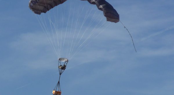 Airborne Systems - FireFly Army Cargo Delivery Parachute System. JPADS 2K System of Choice. Carries unmanned loads up to 2,200 lbs. Max deployment altitude 24,500 ft. Military cargo and inflated canopy, blue sky.