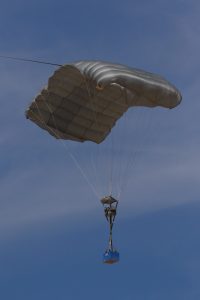 Airborne Systems MicroFly II Army Cargo delivery system. JPADS / GPADS: Guided Precision MilitaryAerial Delivery System. Use with any Airborne Systems Ram Air Canopy. Canopy flying with blue cargo box and blue sky.