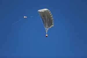Airborne Systems MicroFly II Army Cargo delivery system. JPADS / GPADS: Guided Precision MilitaryAerial Delivery System. Use with any Airborne Systems Ram Air Canopy. Canopy flying with orange cargo box and blue sky.