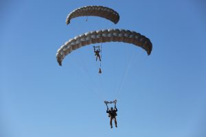 Airborne Systems - MMS Multi-mission Army Ram Air Parachute system for military special forces jumpers. Carries 450-485 lbs. Max deployment altitude 25,000 ft. 2 Jumpers and Ram Air canopies, blue sky.