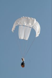 Airborne Systems Unicross Army cargo delivery parachute system. GPADS / JPADS. Low cost, modular design for one-time use or quick repack. Three sizes carry payloads of 75 lbs to 3,200 lbs. White canopy with cargo and blue sky.