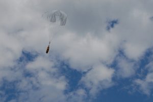 Airborne Systems Unicross Army / military cargo delivery parachute system. Low cost, modular design for one-time use or quick repack. Three sizes carry payloads of 75 lbs to 3,200 lbs. Canopy and orange cargo bags flying with blue sky and clouds at a distance.