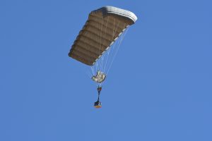 Airborne Systems - FlyClops Army Cargo delivery parachute: one-time use GPADS / JPADS. Carries payloads from 750-2200 lbs. Max deployment altitude 17,500 ft. Military cargo and canopy, blue sky.