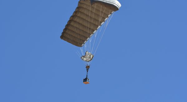 Airborne Systems - FlyClops Army Cargo delivery parachute: one-time use GPADS / JPADS. Carries payloads from 750-2200 lbs. Max deployment altitude 17,500 ft. Military cargo and canopy, blue sky.