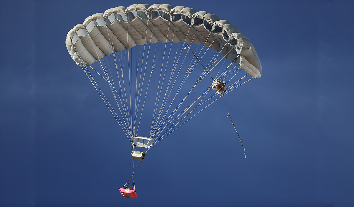 Airborne Systems MicroFly II Army Cargo delivery system. JPADS / GPADS: Guided Precision Aerial Delivery System. Use with any Airborne Systems Ram Air Canopy. Deployed parachute with orange cargo platform and blue sky.