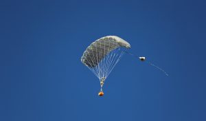 Airborne Systems MicroFly II Army Cargo delivery system. JPADS / GPADS: Guided Precision Aerial Delivery System. Use with any Airborne Systems Ram Air Canopy. Deployed parachute with orange cargo box and blue sky.