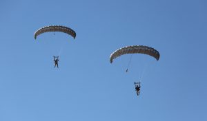Airborne Systems MicroFly II Army Cargo delivery system. JPADS / GPADS: Guided Precision Aerial Delivery System. Use with any Airborne Systems Ram Air Canopy. Two deployed parachutes with military jumpers and blue sky.