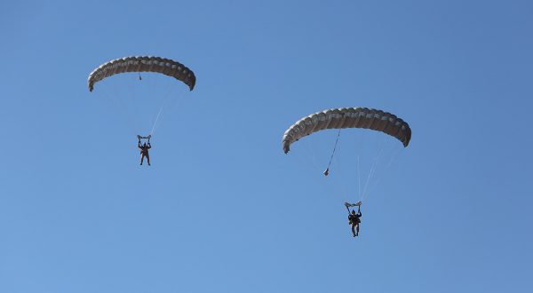 Airborne Systems MicroFly II Army Cargo delivery system. JPADS / GPADS: Guided Precision Aerial Delivery System. Use with any Airborne Systems Ram Air Canopy. Two deployed parachutes with military jumpers and blue sky.
