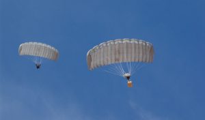 Airborne Systems MicroFly II Army Cargo delivery system. JPADS / GPADS: Guided Precision Aerial Delivery System. Use with any Airborne Systems Ram Air Canopy. Two deployed parachutes from below with one jumper and one white cargo box and blue sky.