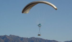 Airborne Systems - DragonFly Army Cargo Delivery Parachute System. JPADS 10K System of Choice. Eliptical canopy carries loads up to 10,000 lbs. Max altitude 24,500 ft. Military cargo on platform and canopy, blue sky and mountains.