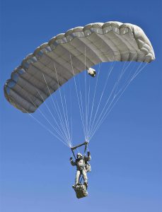 Airborne Systems Intruder RA-1 Army Ram Air Parachute system for military special forces and beginner jumpers. Carries 450 lbs. Max deployment altitude 25,000 ft. Deployed canopy with military jumper / gear bag and blue sky.