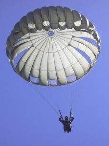 Airborne Systems MC-6 Army Troop Parachute non-steerable for military jumpers. Low opening. Carries up to 400 lbs. Minimum deployment altitude 500 ft. Canopy and jumper from below close up with blue sky.