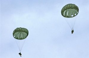 Airborne Systems MC-6 Army Troop Parachute non-steerable for military jumpers. Low opening. Carries up to 400 lbs. Minimum deployment altitude 500 ft. Two deployed canopies and jumpers from a distance with blue sky and light clouds.