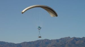 Airborne Systems DragonFly Army Cargo Delivery Parachute System flying with cargo. JPADS 10K System of Choice. Eliptical canopy carries loads up to 10,000 lbs. Max altitude 24,500 ft. View of canopy and payload from the side with mountains behind.