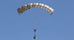 Airborne Systems Canopy with payload. FC Mini Army cargo delivery parachute system. JPADS / GPADS: Guided Precision Aerial Delivery System. Carries 200-500 lbs. Max deployment altitude 24,500 ft. Deployed canopy with green cargo box with blue sky.