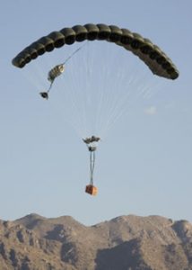 Airborne Systems FireFly Army Cargo Delivery Parachute System. JPADS 2K System of Choice. Carries unmanned loads up to 2,200 lbs. Max deployment altitude 24,500 ft. mountains. Deployed canopy with orange cargo box, mountains and blue sky.