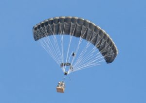 Airborne Systems FireFly Army Cargo Delivery Parachute System. JPADS 2K System of Choice. Carries unmanned loads up to 2,200 lbs. Max deployment altitude 24,500 ft. Deployed canopy with cargo box and blue sky.