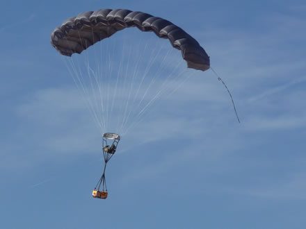Airborne Systems FireFly Army Cargo Delivery Parachute System. JPADS 2K System of Choice. Carries unmanned loads up to 2,200 lbs. Max deployment altitude 24,500 ft. drop. Deployed canopy with orange cargo box, blue sky and whispy clouds.