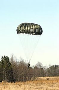 Airborne Systems MC-6 Army Troop Parachute non-steerable for military jumpers. Low opening. Carries up to 400 lbs. Minimum deployment altitude 500 ft. Soldier landing near forest with green canopy.