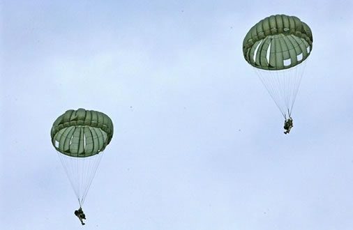 Airborne Systems MC-6 Army Troop Parachute non-steerable for military jumpers. Low opening. Carries up to 400 lbs. Minimum deployment altitude 500 ft. Two jumpers with green parachutes blue sky.