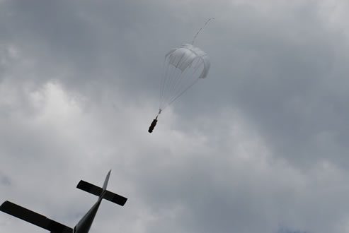 Airborne Systems. Unicross Army cargo delivery parachute system. Low cost, modular design for one-time use or quick repack. Three sizes carry payloads of 75 lbs to 3,200 lbs. Aircraft tail trailed by unicross and cargo with cloudy sky.