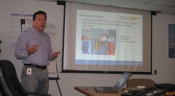 Vincent Mignot, International Business Unit Manager at Airborne Systems teaching class for Camden Youth Aviation Program. Screen behind him reads, "Emergency Escape; Airborne Systems. A leading provider of..." Military Parachute Training. Troop and cargo parachute certification for army riggers & jumpers. Instruction for oxygen and JPADS systems.