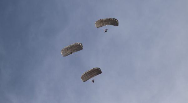Airborne Systems Hi-5 Army Ram Air Parachute Personnel product system for military special forces jumpers with glide modulation. Carries 485 lbs. Max deployment altitude 25,000 feet. Deployed canopy and 3 parachutists from below with blue sky.